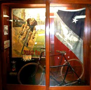 Museum display case with a Frank Kramer Pierce Racer poster, an eagle banner and a Pierce track racing bicycle
