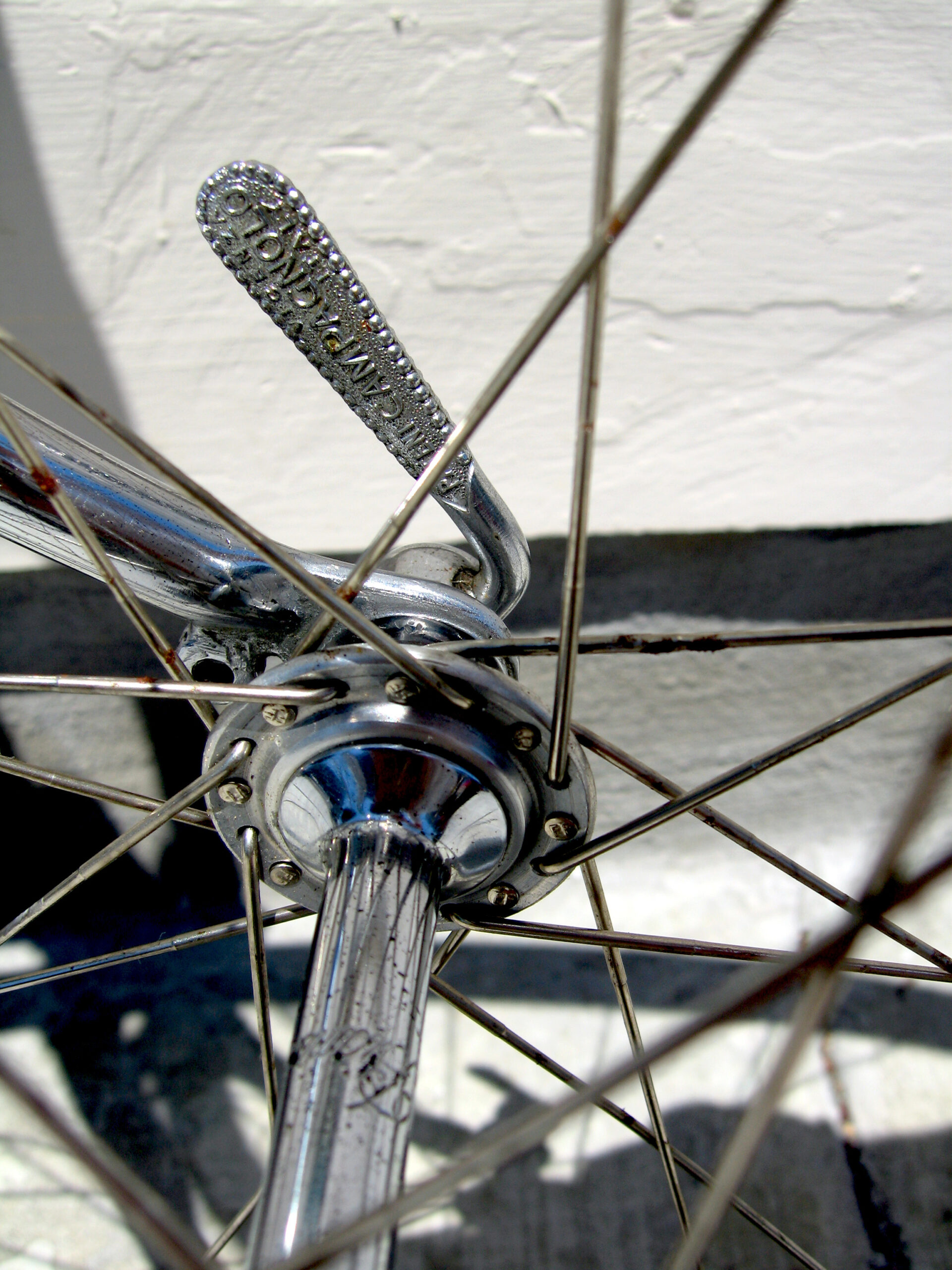 Close view of a chromed quick-release lever on a front hub, the lever face stippled inside a beaded border and lettered PATENT CAMPAGNOLO, seen through the spokes against a white wall