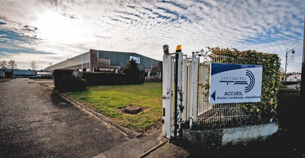 Present-day photograph of the Specialites TA factory entrance gate under a wide sky