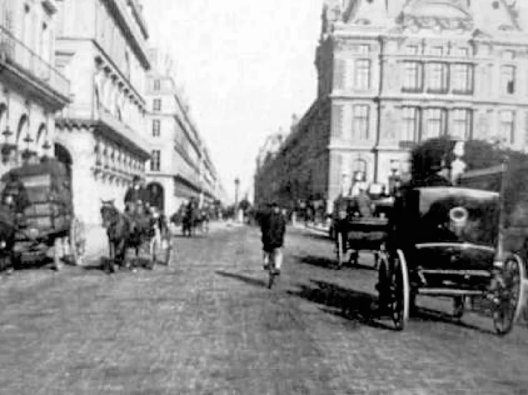 A wide Paris boulevard about 1890, lined with Haussmann facades, where a lone cyclist rides away between horse drawn cabs and delivery wagons