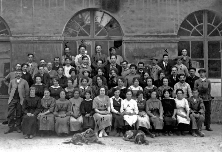 Group portrait of about sixty Tron et Cie workers in 1922, men and women ranged in rows outside the arched factory windows with two dogs lying at the front