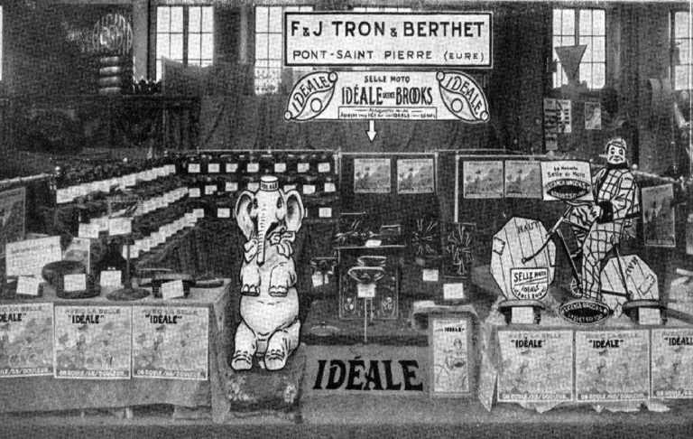 The F & J Tron et Berthet stand at the 1929 Salon du Cycle in Paris, banked with saddles under a Selle Moto Idéale avec Brooks banner, a giant cardboard elephant and a clown figure among the display cards