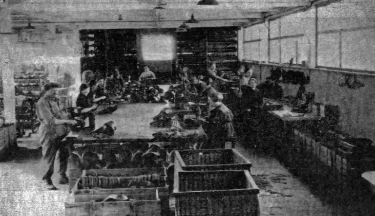 The Idéale assembly shop in 1935, workers ranged along a long bench with finished saddles stacked in wicker crates in the foreground and racks of parts behind