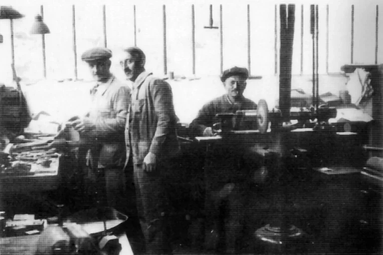 Three men of F. Tron et Cie at a workshop bench beside a lathe, photographed against a wall of factory windows