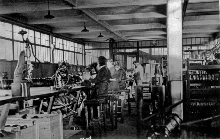 The leather assembly shop at Idéale, women working at a long bench among saddle frames and coiled springs, with racks of springs stacked at right under a wall clock