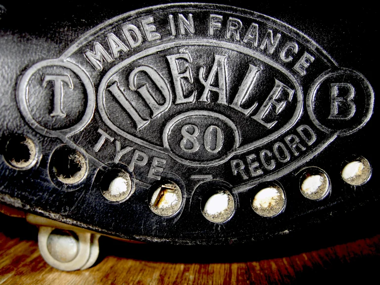 Macro of the embossed Idéale Type 80 Record badge on black leather, Made in France arched above the oval between the T and B roundels, with copper rivets along the flap