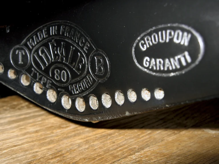 Wide macro of a black Idéale flap carrying both the Type 80 Record badge and the Croupon Garanti oval, its skirt of silvered rivets running above an oak bench