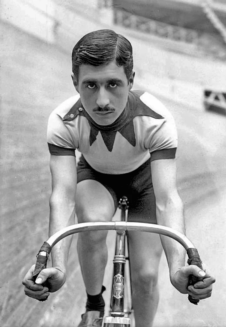 Head-on portrait of Marcel Berthet on a track bicycle at the velodrome, in a chevroned jersey with both hands on the bars