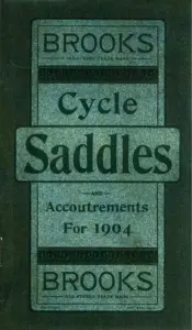 Catalog cover printed in dark green on a mottled green ground. A ruled panel holds the word BROOKS in outline capitals above the line REGISTERED TRADE MARK, then a band of stylised foliate ornament, then the word Cycle in a script face. A raised rectangular cartouche across the middle carries the word Saddles in large bold letters. Below it the words AND, Accoutrements and For 1904 are set in three lines, followed by a second band of ornament and the word BROOKS repeated in outline capitals above REGISTERED TRADE MARK. The word COPYRIGHT is printed at the lower left of the panel and ENT. STA. HALL. at the lower right.