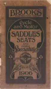 Catalog cover printed in dark brown on a tan card, worn at the edges and with a hole punched at the upper left. A ruled border encloses a panel with the word BROOKS in heavy shaded capitals above REGISTERED TRADE MARK, then the words Cycle and Motor in a flowing script, then SADDLES and SEATS in bold capitals within an arched cartouche, then AND OTHER above the word Specialities in a large italic script with a long flourish. Below sits a roundel of a bee with outspread wings encircled by the word INDUSTRIA and flanked by scrolls lettered TRADE and MARK, and beneath that a scrolled banner reading FOR above the year 1906. COPYRIGHT is printed at the lower left of the panel and ENT. STA. HALL. at the lower right.