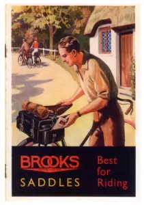 Color lithograph of a young man in shirtsleeves beside his bicycle outside a thatched cottage in autumn, sliding a map into a large black saddlebag with a rolled cape strapped above it, two more riders freewheeling away down the lane behind him; across the foot a black band reads BROOKS SADDLES in red and yellow with Best for Riding at the right.