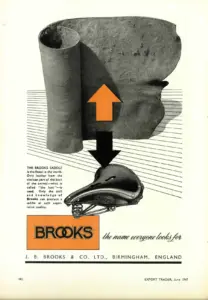 Full-page advertisement in black and orange on cream. A large photographic detail of a thick rolled hide fills the upper half, with an orange and black arrow leading down from it to a photograph of a finished sprung leather saddle. A block of small text runs down the left margin and a solid orange panel at the foot carries the Brooks name beside the line the name everyone looks for.