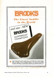 Full-page advertisement in black and orange on white within a fine rule border. A colour photograph of a plain unsprung honey-brown sports saddle sits in the centre against a black flash, with the words and now NEW above it and BROOKS LIGHTWEIGHT CHAMPIONS in outlined capitals beneath. A paragraph of small type at the foot explains the range, above the firm's name and Birmingham address.