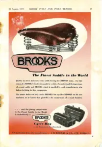 Full-page advertisement whose upper two-thirds is a deep orange ground carrying a large photograph of a fully sprung tan leather saddle within a white ellipse, the Brooks name below it in outlined capitals. The lower third is white and carries three paragraphs of text, with a small photograph of two rectangular cycle bags at the foot beside the Brooks name plate.