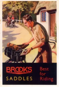 Full-color printed advertisement showing a young man in an open-necked shirt standing over a bicycle outside a thatched cottage, tucking a folded map into a large black saddlebag with a tan bedroll strapped above it, two loaded cyclists riding away up the lane behind him, and a black band across the foot carrying BROOKS in red with SADDLES and the words Best for Riding.