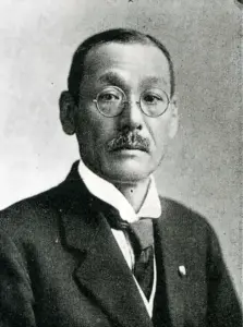 Half-length studio portrait of Okazaki Kyujiro, a middle-aged man in a dark suit, wing collar and round wire-rimmed spectacles, photographed about 1929