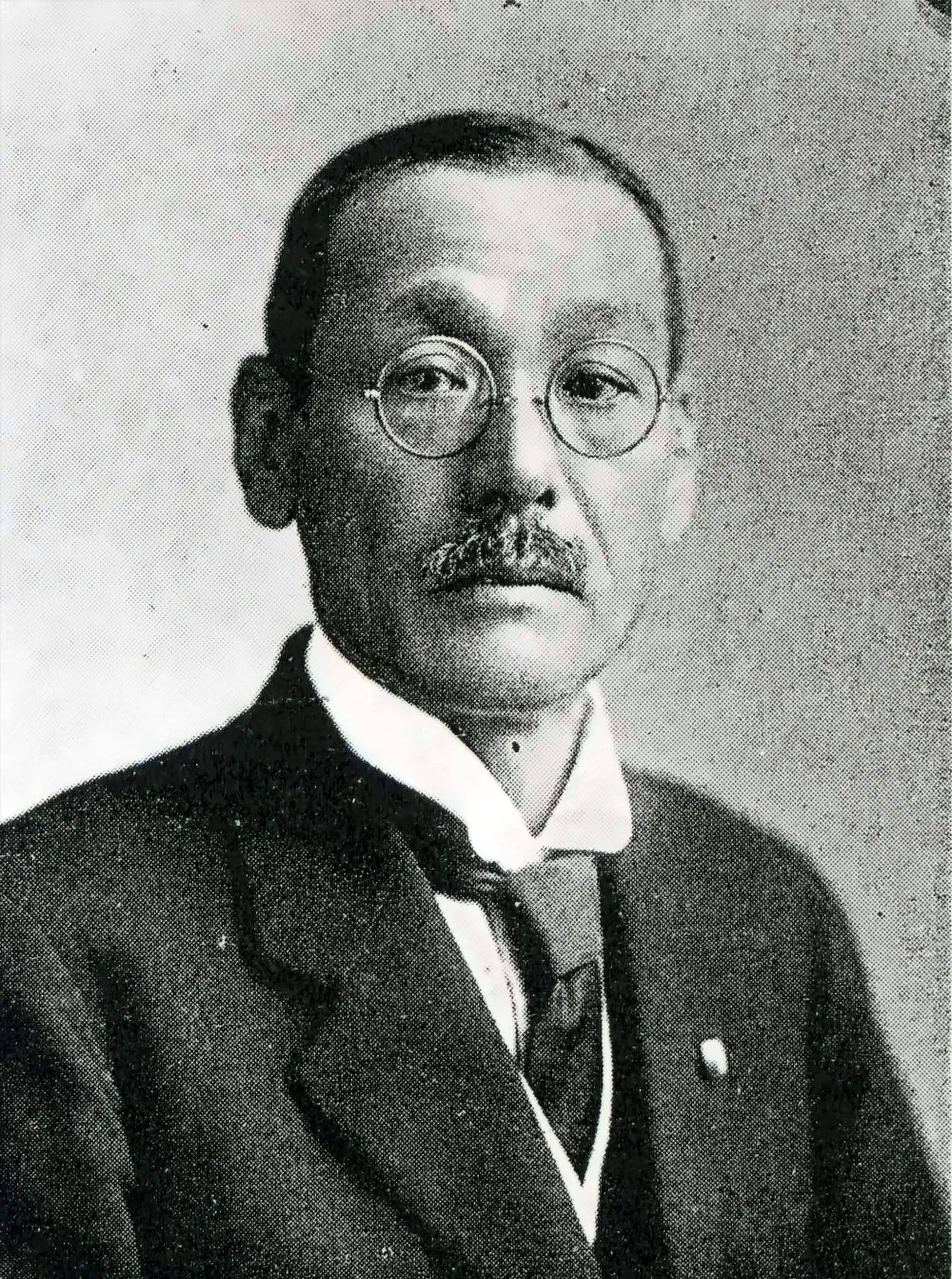 Half-length studio portrait of Okazaki Kyujiro, a middle-aged man in a dark suit, wing collar and round wire-rimmed spectacles, photographed about 1929