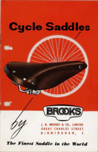 A brown Brooks leather saddle, its side panel stamped "BROOKS" and "B17", pictured against a wheel drawn in white on a red ground, with the BROOKS logo panel and the company address on the white band below.