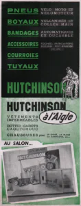Hutchinson advertisement from L'Officiel du cycle, 23 November 1957, page 39: a tall two-color panel: on a black field, green capitals list the firm's rubber products, with a cartoon of a stout mustached man in a small hat and polka-dot overalls straddling a stool as he spins a bicycle wheel, and below, on a green field, a photograph of the Hutchinson stand at the Salon, with tires coiled along the floor, wheel and tire displays on the back wall, a large cutout of the same cartoon figure and a row of chairs.