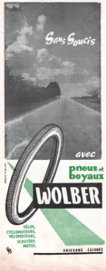Wolber advertisement from L'Officiel du cycle, 3-17 August 1959, page 2: a tall photograph of an empty country road running straight to the horizon under a cloudy sky, lined with trees on the left, with a drawn tire rolling up out of the foreground and a green dotted band sweeping across the lower half; the name Wolber is reversed out in white on a black panel.