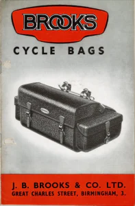 A large black grained Brooks cycle bag with an end pocket, buckled straps and a rod with two saddle clips across its top, shown in a photograph on a gray ground under the red BROOKS logo panel, with the company name and address in a red band at the foot.