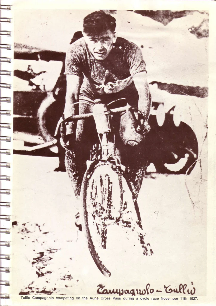 A sepia photograph of Tullio Campagnolo riding head-on toward the camera through snow and slush, mud-spattered, with spare tubulars over his shoulders and a car behind him, printed full page in Campagnolo's 1974 catalog.