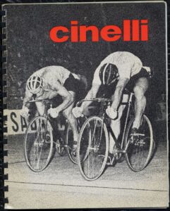 Front cover of the Cinelli catalog: the name "cinelli" in large red lowercase letters across the top of a coarse-grained black-and-white photograph of two track sprinters bent low over their bicycles side by side on a velodrome, with part of a trackside board lettered "SA" behind them.